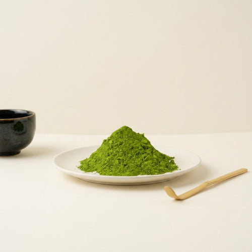 A mound of bright green matcha powder sits on a small white plate, with a dark ceramic cup at left and a wooden scoop at right on a clean off-white surface.